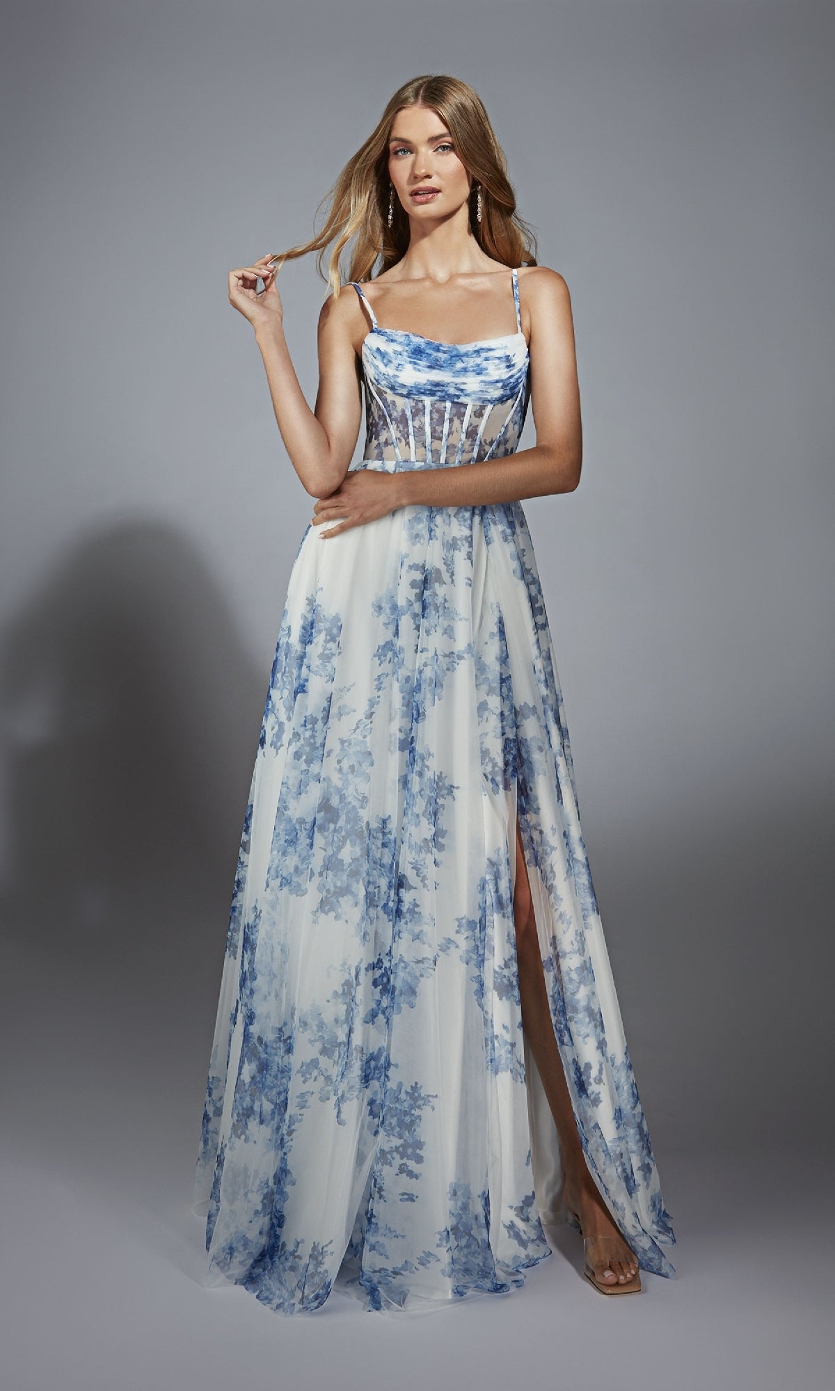 Woman in the Alyce 61752 long prom dress, featuring a white and blue floral pattern, cowl neckline, spaghetti straps, and thigh-high slit, standing against a plain gray background.